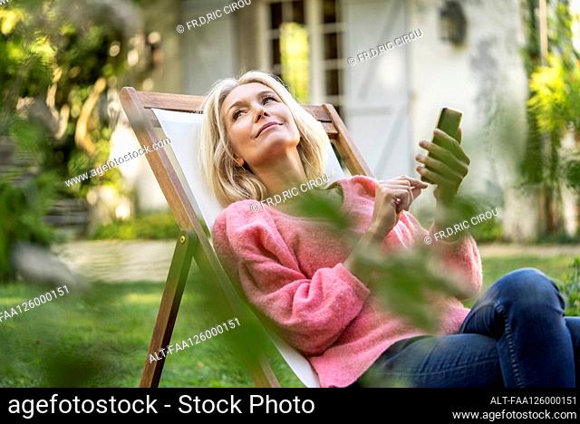 Thoughtful mature woman using smartphone while sitting on deckchair