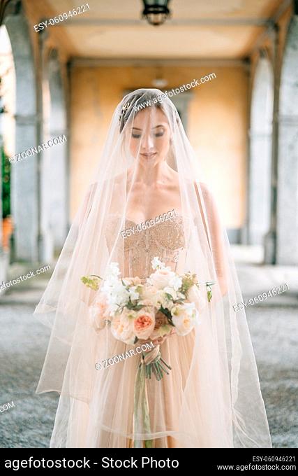 Bride in a wedding dress and a veil with a bouquet stands with her eyes downcast on an old terrace with columns. Lake Como. High quality photo