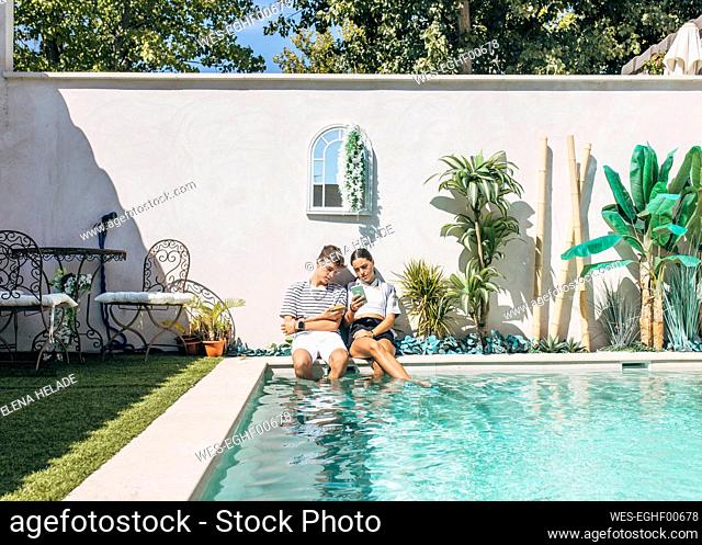 Teenage boy with sister using smart phone sitting by swimming pool on sunny day