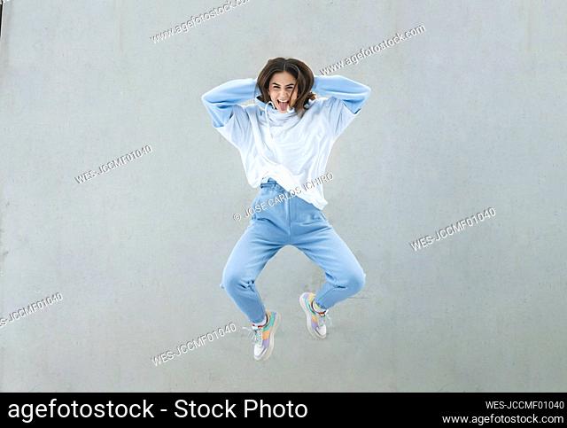 Carefree woman with hands behind head jumping against gray wall