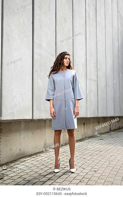 Cute girl in the gray jacket stands on the background of the concrete wall. She has white-blue shoes. She looks to the right. Outdoors. Vertical