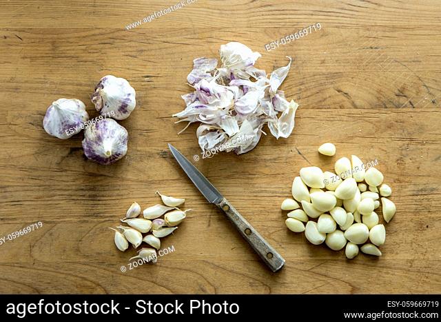 An overview of garlic bulbs, cloves, and peels and a knife on a wooden cutting board