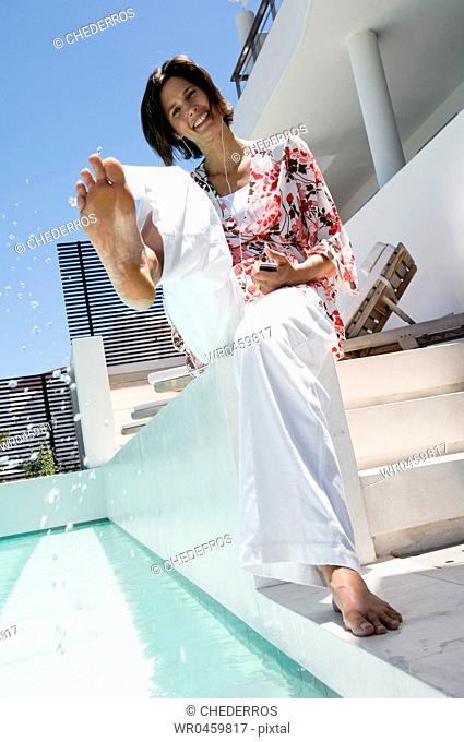 Portrait of a mid adult woman splashing water at the poolside and listening to an MP3 player