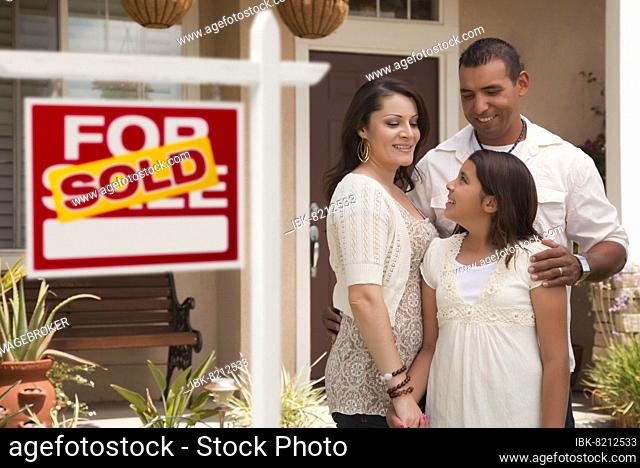 Hispanic mother, father and daughter in front of their new home with sold home for sale real estate sign