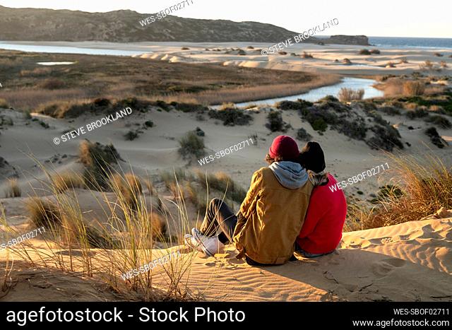 Couple wearing knit hat sitting together on sand dune during sunset