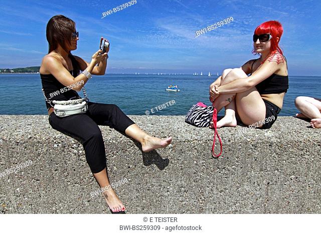 two young woman sitting on quay wall, taking photos from each other, Germany, Schleswig-Holstein, Travemuende, Luebeck