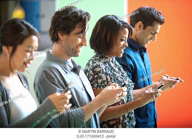 Smiling men and women text messaging on cell phones in a row