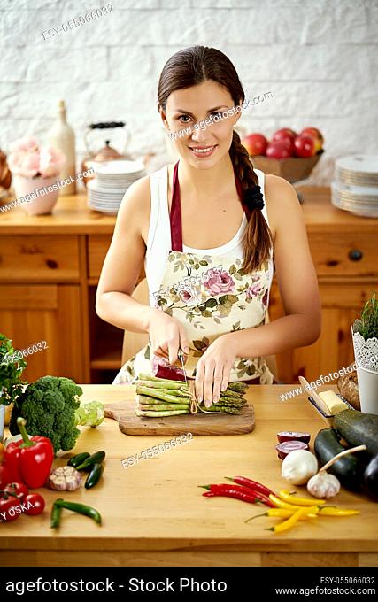 beautiful young woman, brunette slicing asparagus in the kitchen at a table full of organic vegetables