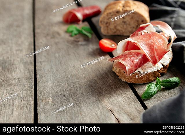 Italian cuisine concept. Crusty bread toast with cream cheese and thinly sliced ham, cherry tomatos, basil leaves branches, kitchen towel on wooden background