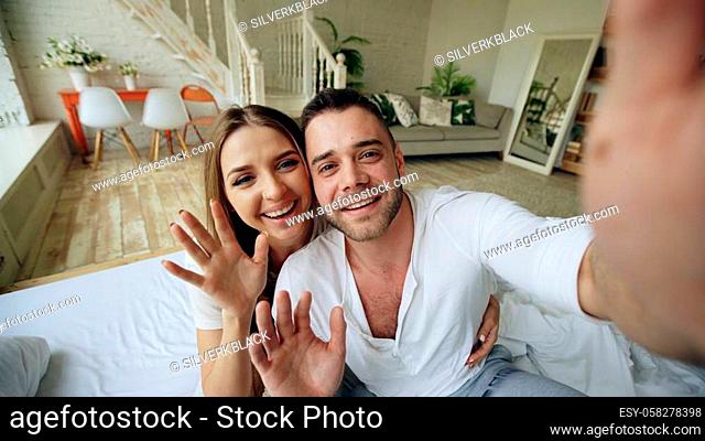 Young cute and loving couple having video chat holding tablet computer camera and chatting to friends sitting in bed at home