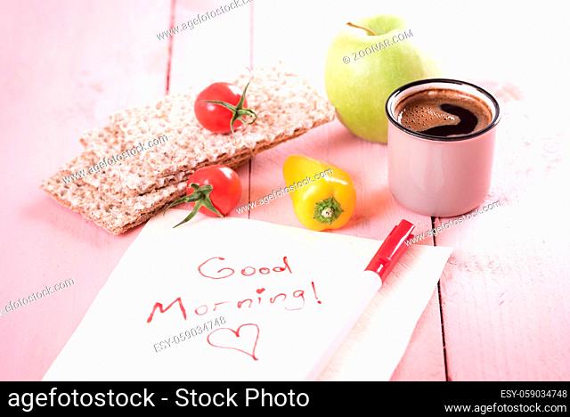 Healthy snack with whole-wheat crispbread, vegetables, apple, a cup of coffee and a napkin with a good morning message on a pink background, in daylight