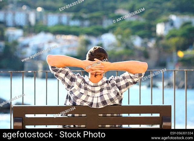 Back view portrait of a man relaxing sitting on a bench contemplating ocean in a coast town
