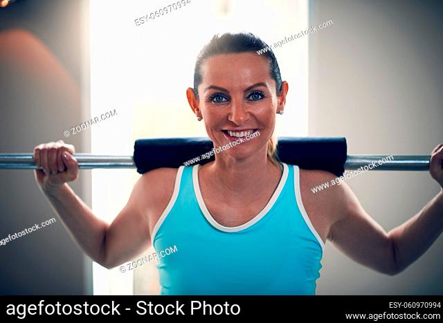 Attractive blonde woman in mint sleeveless shirt standing in the gym, holding barbell on her shoulders and looking at camera