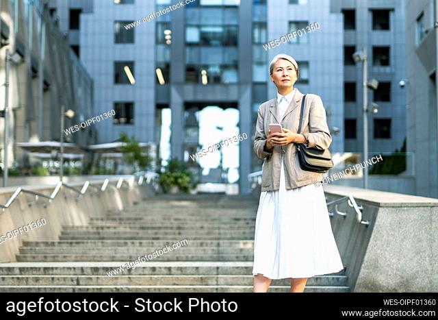 Mature woman with smart phone standing on stairs in city