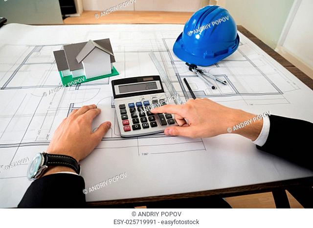 Close-up Of Architect With Blueprint Calculating On Calculator At Desk