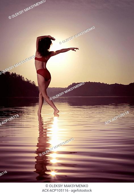 Young woman in swimsuit dancing in the sun on the water in beautiful morning sunrise scenery. Muskoka, Ontario, Canada