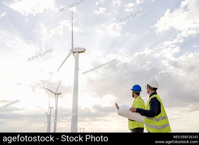 Technicians looking at wind turbines by clouds in sky