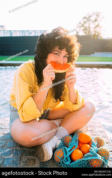 Woman with oranges in mesh bag eating watermelon by pond