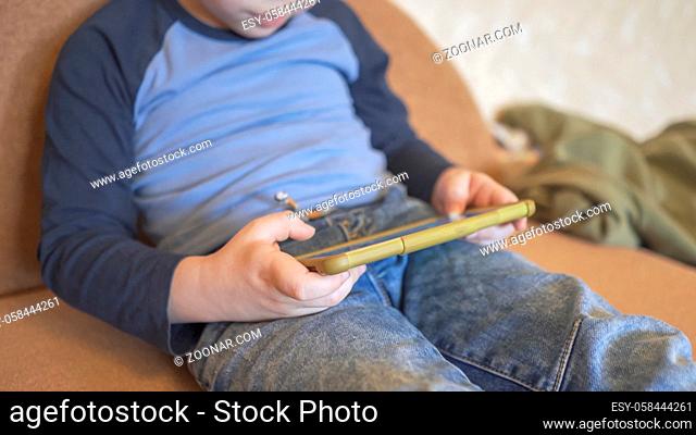 Boy Uses Phone Sitting On The Couch, Caucasian Boy In Casual Wear Spends Time Playing Or Learning Over Phone At Home, Little Boy Needs To Stay At Home Due To...