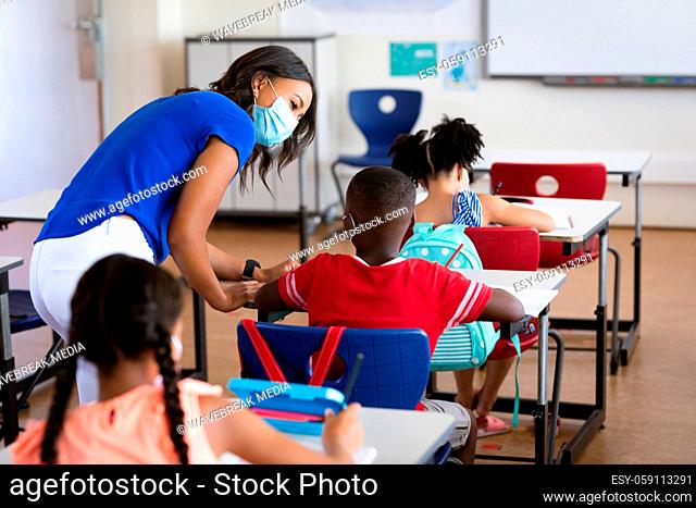 African american female teacher wearing face mask teaching african american boy in class at school