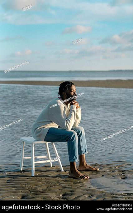 Young man sitting on white stool while watching sunset at beach
