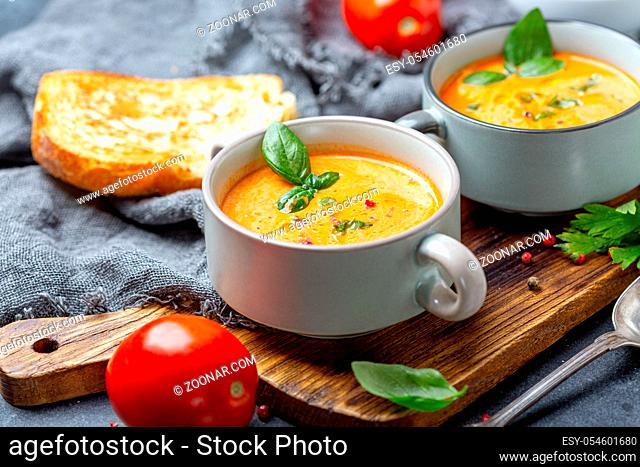 Delicious tomato soup with spices and green basil in ceramic pot on wooden serving board, selective focus