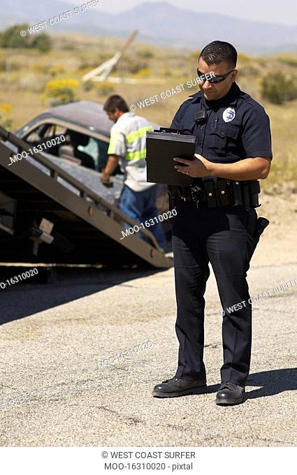 Police officer writing notes tow truck driver lifting crashed car in background