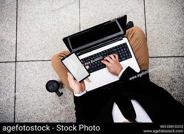 Businessman with mobile phone using laptop while sitting on footpath