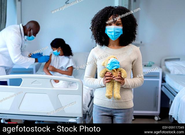 Mixed race woman in face mask holding teddy in hospital doctor giving covid vaccine to sick daughter