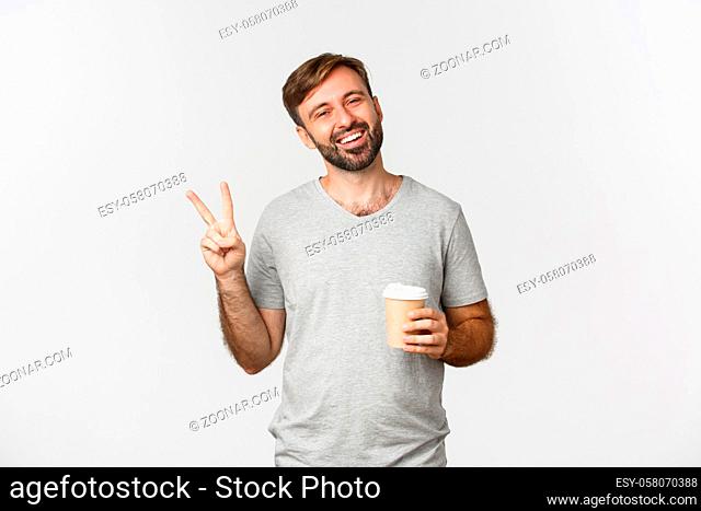 Portrait of carefree smiling man in gray t-shirt, drinking coffee and showing peace sign, standing over white background