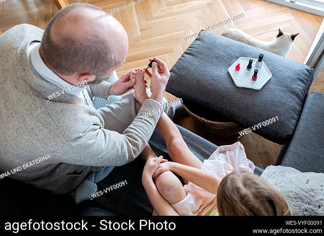 Father painting his daughter's toenails with red nail polish on the couch in living room, top view