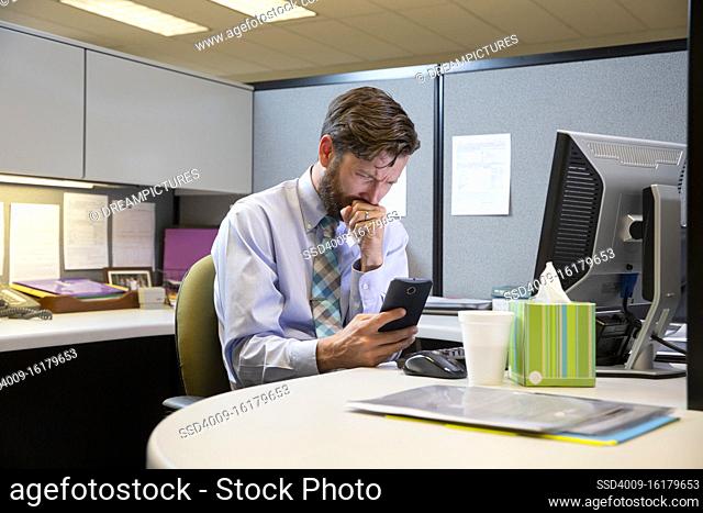 coughing young Caucasian man working in cubicle at office, fighting off a cold with tissues and hot tea, checking cell phone