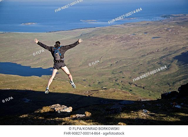 Fell runner jumping over rock (imitating base jump), coming down from mountain peak, with Loch an t-Siob in background, Beinn an Oir, Paps of Jura, Isle of Jura