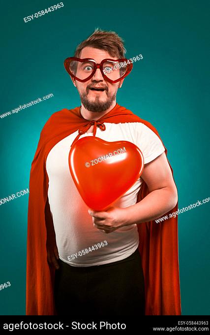 Superman In Love Holds Big Red Heart. Positive Man In Superhero Costume And Heart-Shaped Glasses Poses On Biscay Green Background