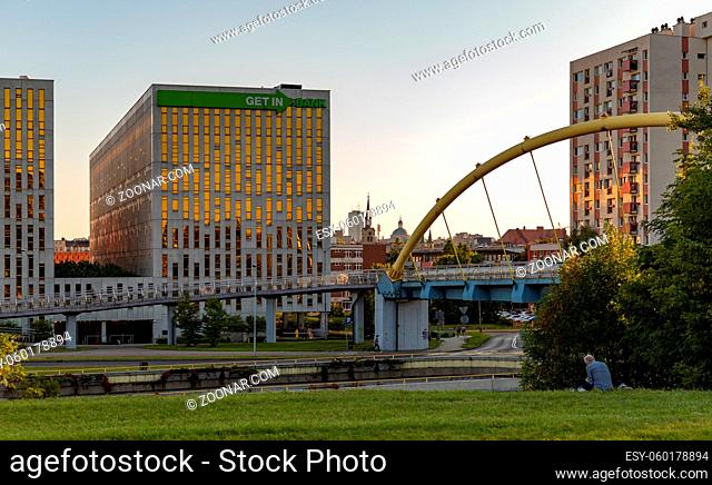 A picture of the Ro?dzie?skiego Avenue, the pedestrian bridge, the buildings around it and a man sitting on the grass