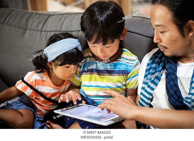 Man, boy and young girl sitting on a grey sofa, looking at digital tablet