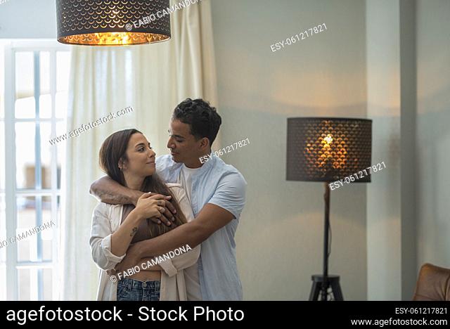 Young millennial interracial couple hug with love at home in living room - relationship with black boy and caucasian girl together standing and embracing...