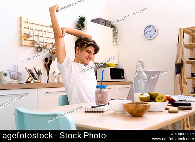 Young man stretching his hand while sitting at table with laptop in kitchen