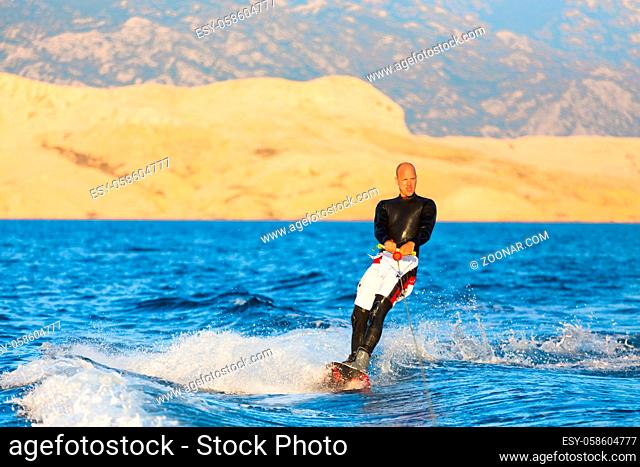 Wakeboarder in wetsuit riding in sunset. Wakeboarding is a surface water sport which involves riding a wakeboard over the surface of a body of water