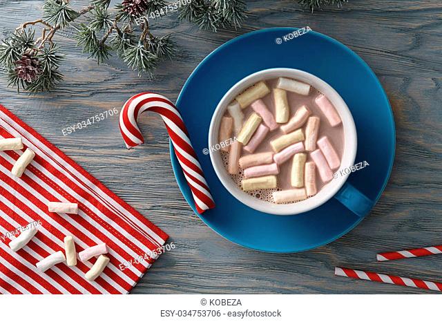 A big cup of hot cocoa with marshmallows decorated with striped cloth, candy cane and fir tree branch on a blue wooden background. Top view