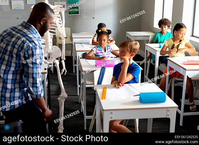 African american young male teacher showing skeleton to multiracial elementary students in class