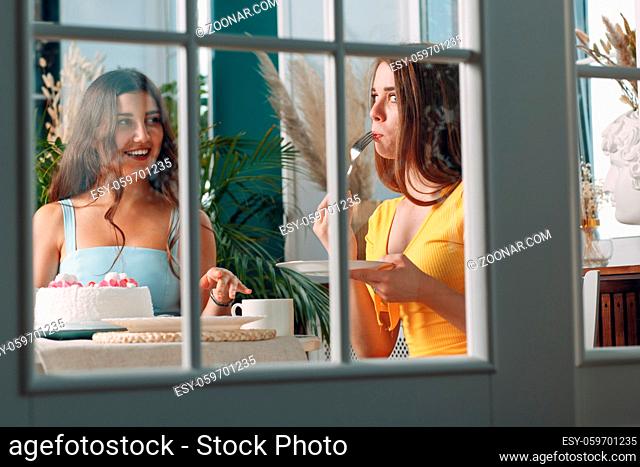 Women friends at home sitting and smiling with white birthday cake behind glass door