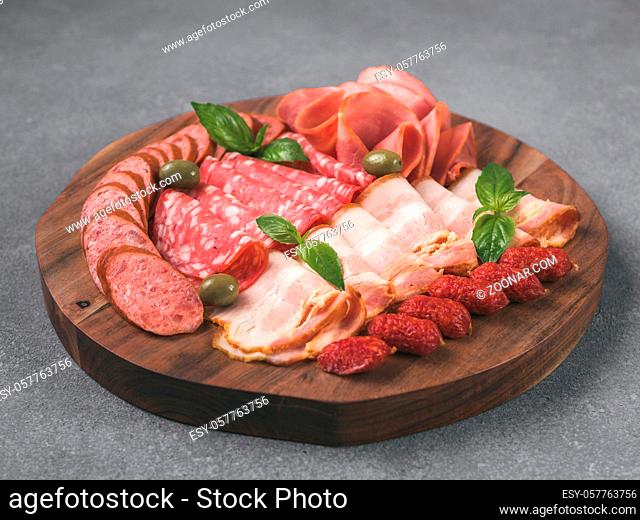 Antipasto set platter on wooden plate over gray stone. Cold smoked meat plate with sausage, sliced ham, prosciutto, bacon, olives and basil