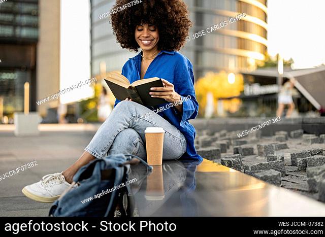 Smiling Afro woman reading book sitting by disposable coffee cup on wall