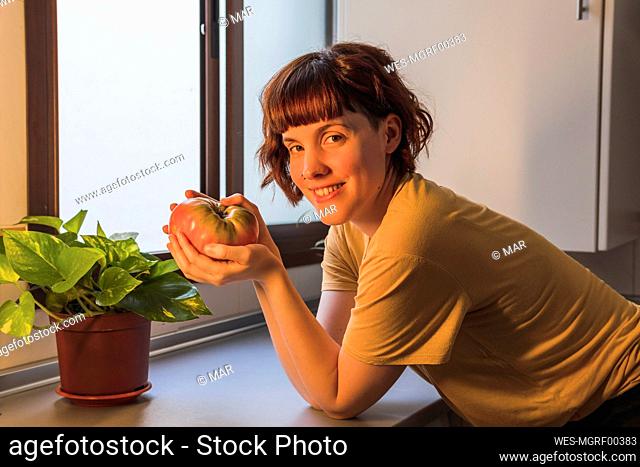 Smiling beautiful woman with beefsteak tomato leaning on kitchen counter at home