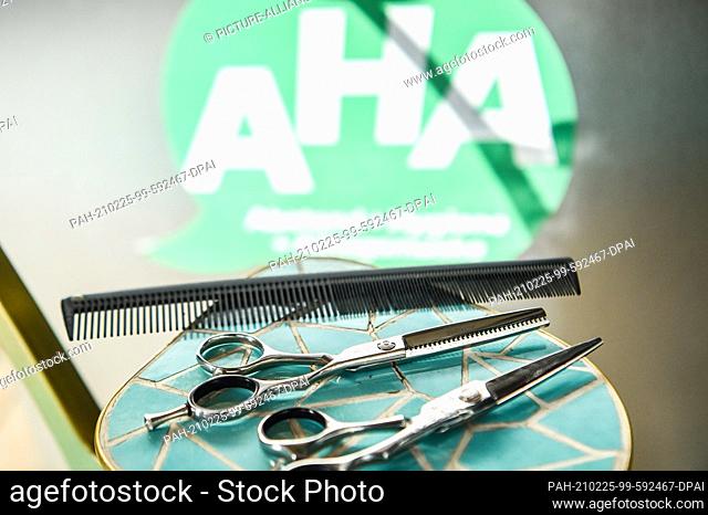 25 February 2021, Berlin: In a closed hairdressing salon, hair scissors and a comb lie on a small table. In the background is a sticker with ""AHA""