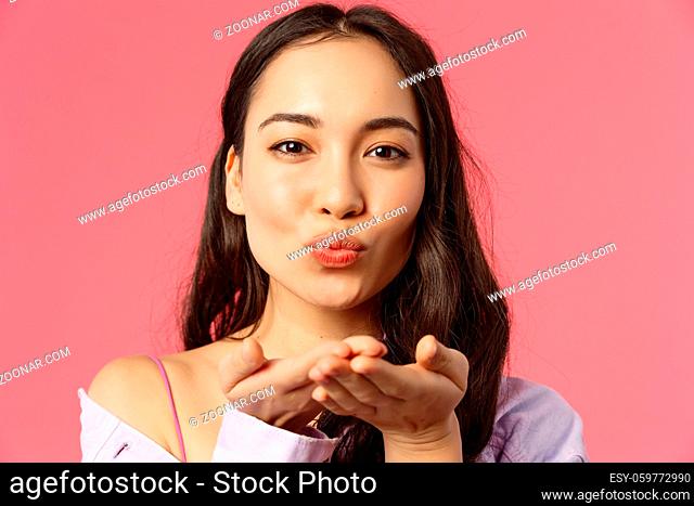 Close-up portrait of tender, romantic and passionate feminine young asian woman, sending air kiss with hand near folded lips, smiling lovely, concept of beauty