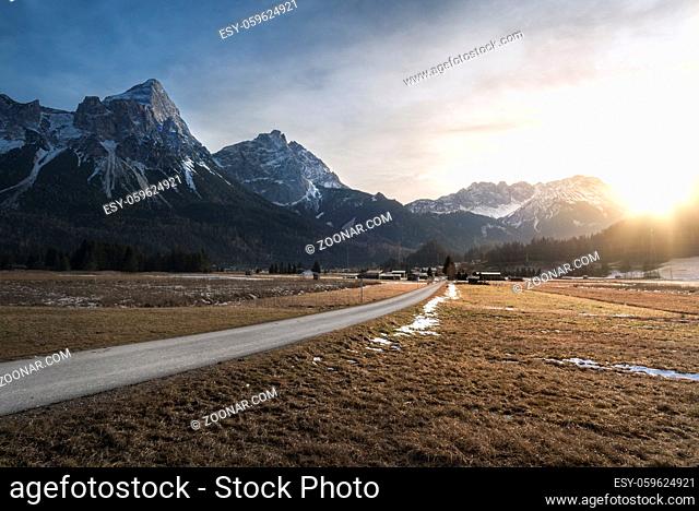 Winter sunset over the snow-capped peaks of the Alps mountains, a country road, a dried grass meadow and a village, near Ehrwald, Austria
