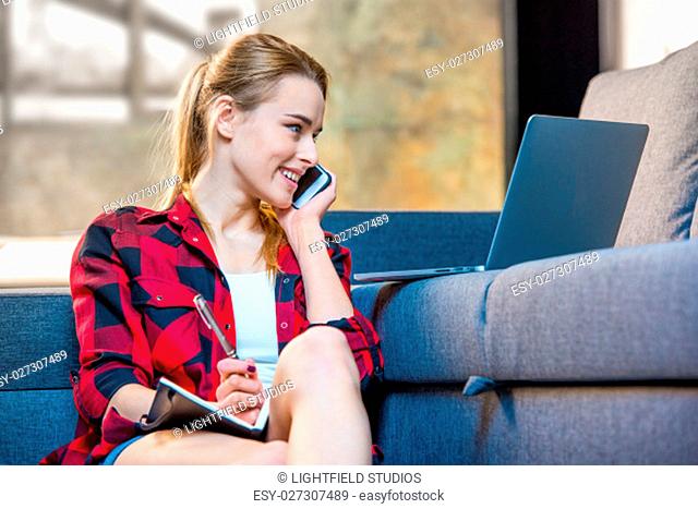 Teenage girl talking on smartphone, making notes in notebook and looking at laptop