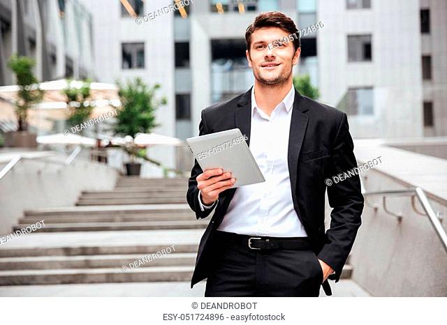 Happy confident young businessman with tablet walking in the city